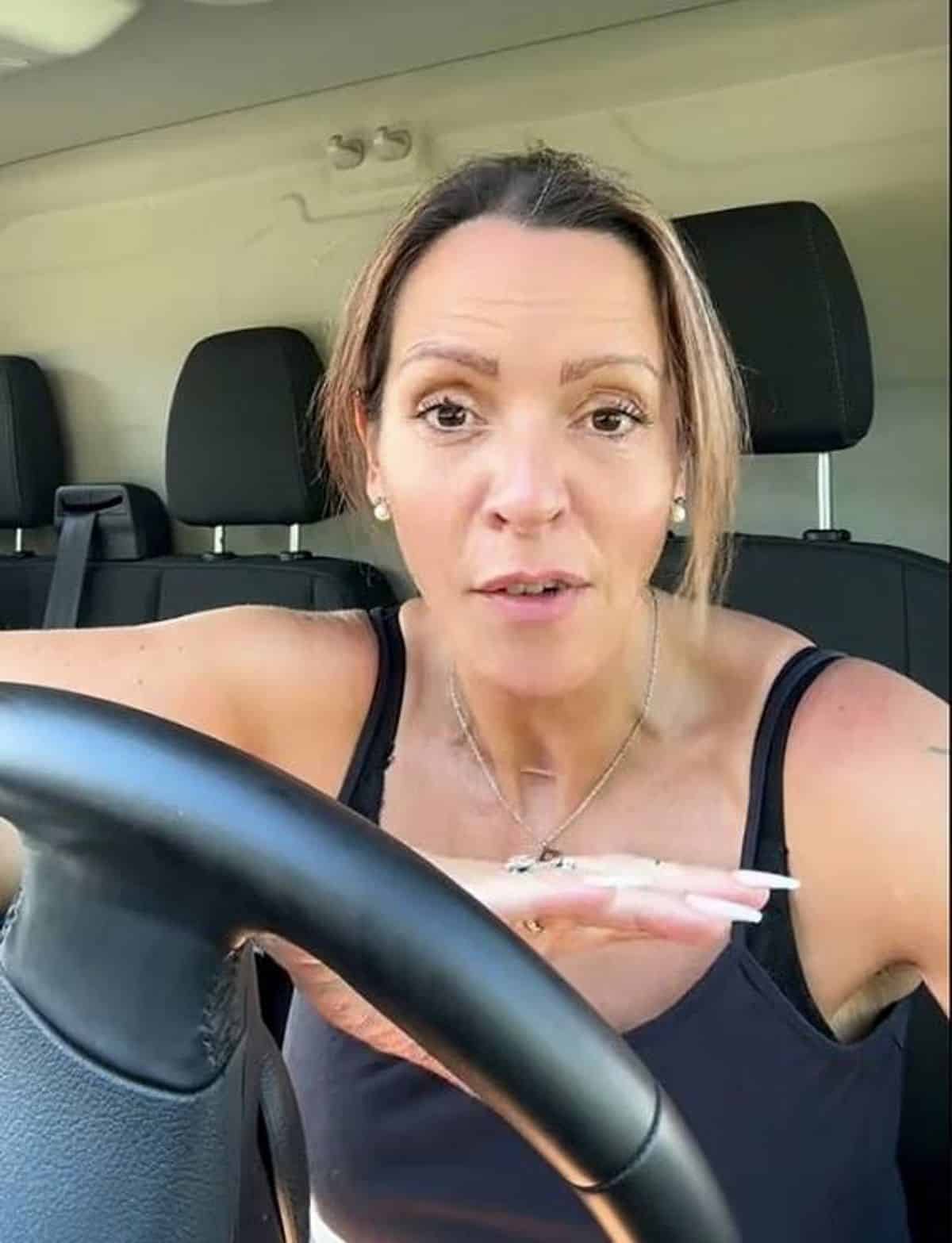 An adult female is seated inside a vehicle, with her hand resting on the steering wheel. She is dressed in a black sleeveless top and appears to be focused on something in front of her. The interior of the vehicle features black seats with visible headrests.