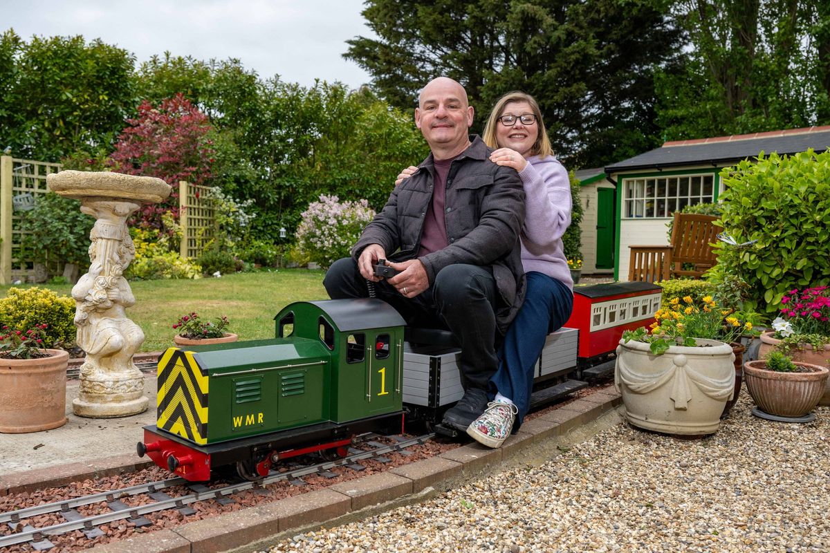 Two individuals are seated on a miniature train, possibly engaged in a recreational activity. The setting is a garden adorned with various plants, trees, and a building in the background. The ground is covered in gravel.