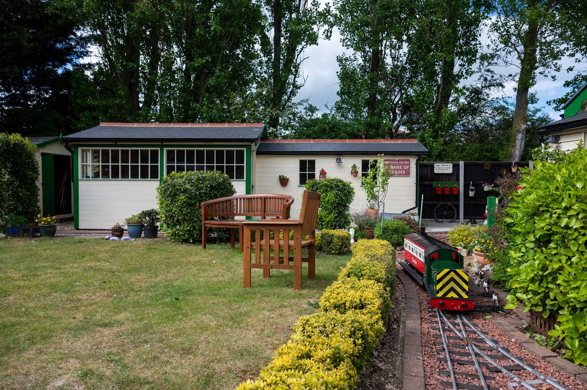 A charming single-story house with a tiled roof is situated in a lush garden, featuring a wooden bench, a table, and a variety of well-maintained plants and shrubs. The property is set against a backdrop of tall trees, creating a serene and picturesque environment.