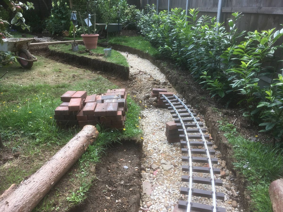 An outdoor area with a gravel pathway resembling a railway track, bordered by neatly arranged garden beds. The track is partially constructed with gravel and a few bricks, and a wooden log is placed nearby, suggesting ongoing construction or landscaping work. Surrounding greenery includes various shrubs and plants, contributing to the natural ambiance of the garden.