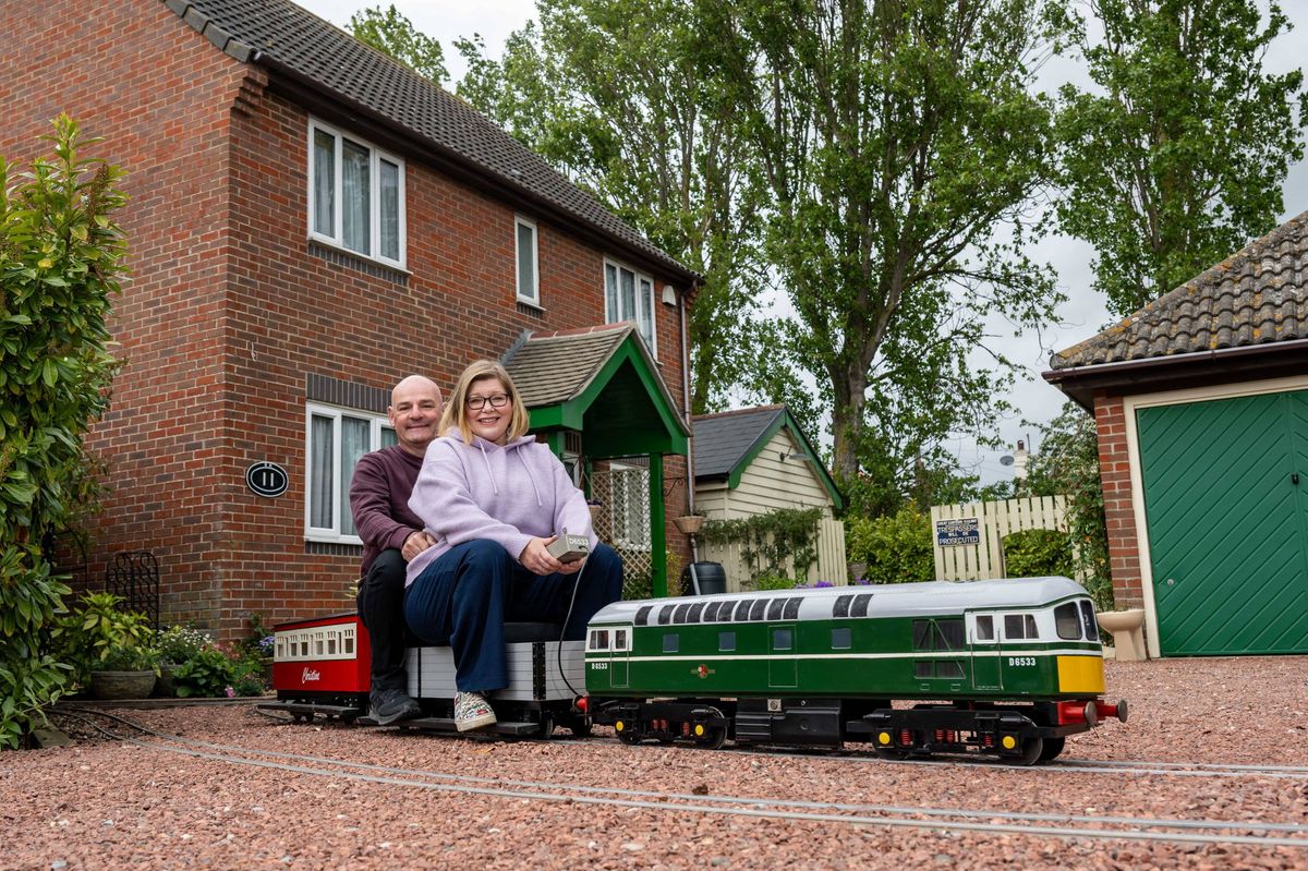 An image depicts two individuals sitting on a small-scale model train positioned in front of a residential building. The setting includes greenery and other houses in the background.