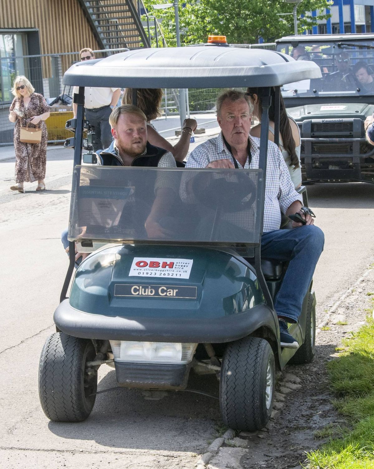Two individuals are seated within a golf cart, navigating the road. A woman with a handbag can be seen walking behind the vehicle. The surroundings include additional vehicles and pedestrians, indicating a bustling street.