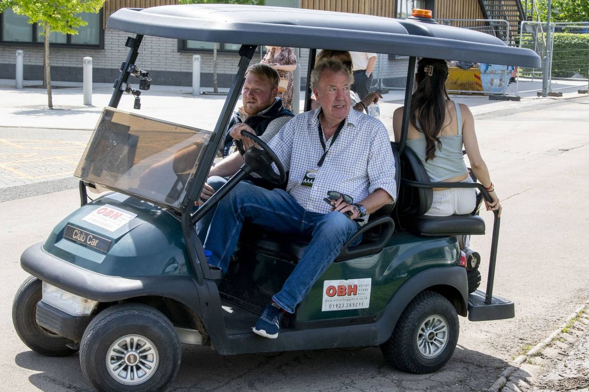 An individual is driving a golf cart with two passengers on a paved road, with a building visible in the background.
